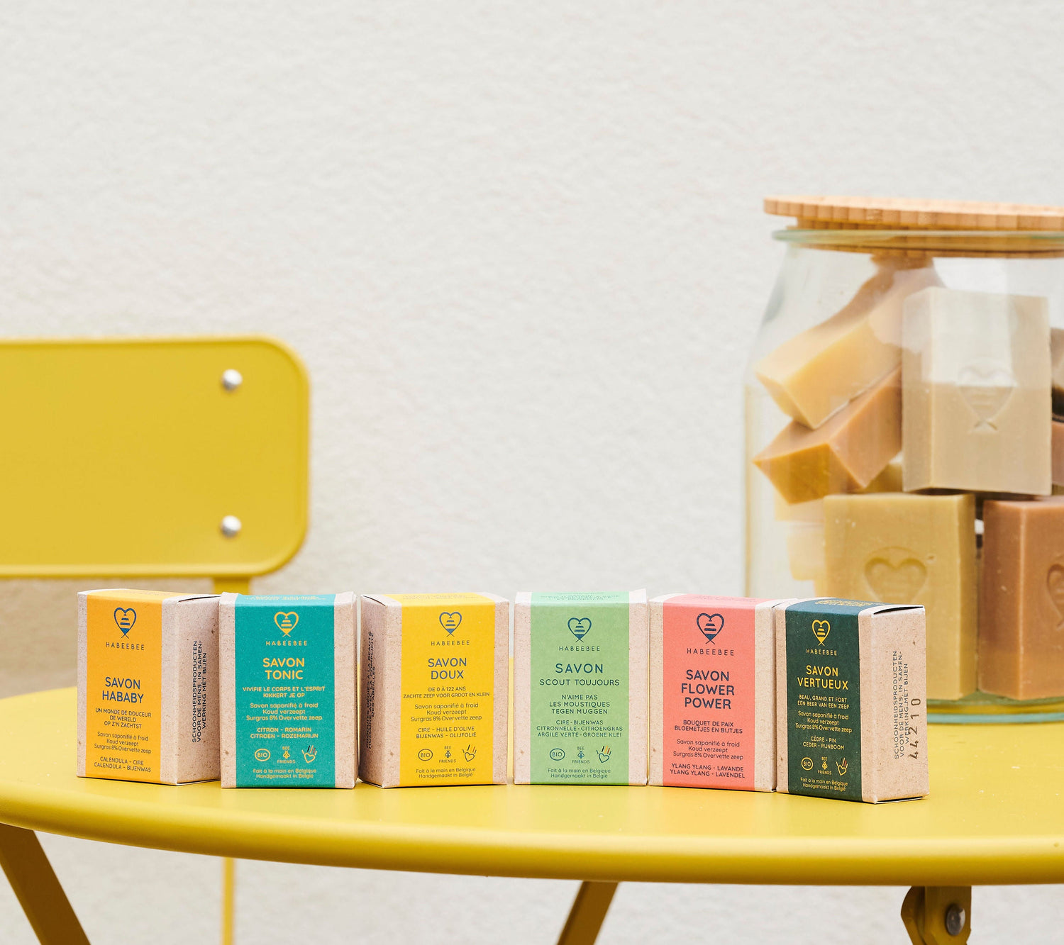 Savons solides Habeebee colorés sur une table jaune avec un bocal rempli de savons en arrière-plan.Kleurrijke vaste zepen van Habeebee op een gele tafel met een pot vol zepen op de achtergrond.Colorful Habeebee solid soaps on a yellow table