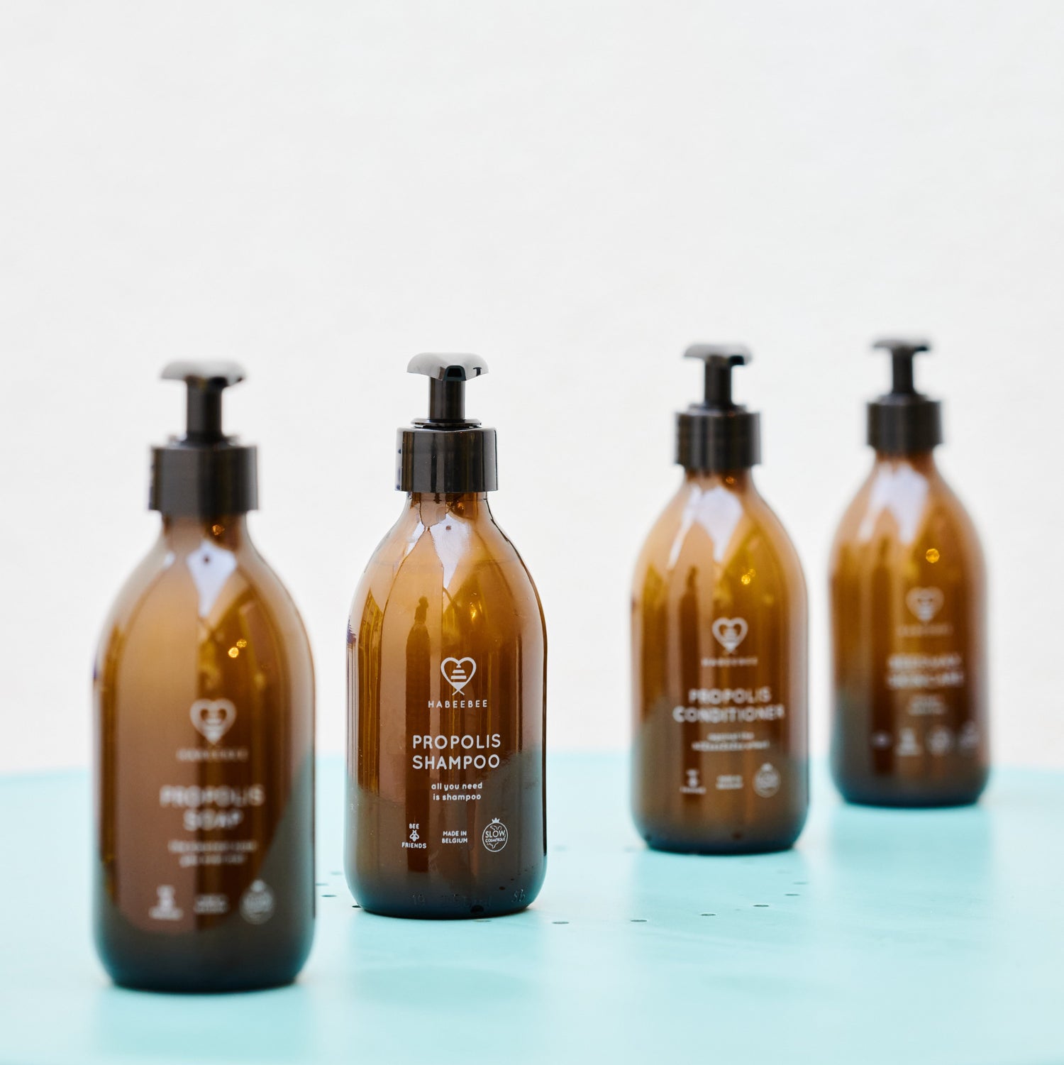 Quatre flacons en verre ambré Habeebee (savon, shampooing, après-shampooing, lait) posés sur une surface bleue. Vier amberkleurige glazen flessen van Habeebee op een blauwe ondergrond (zeep, shampoo, conditioner, lotion).