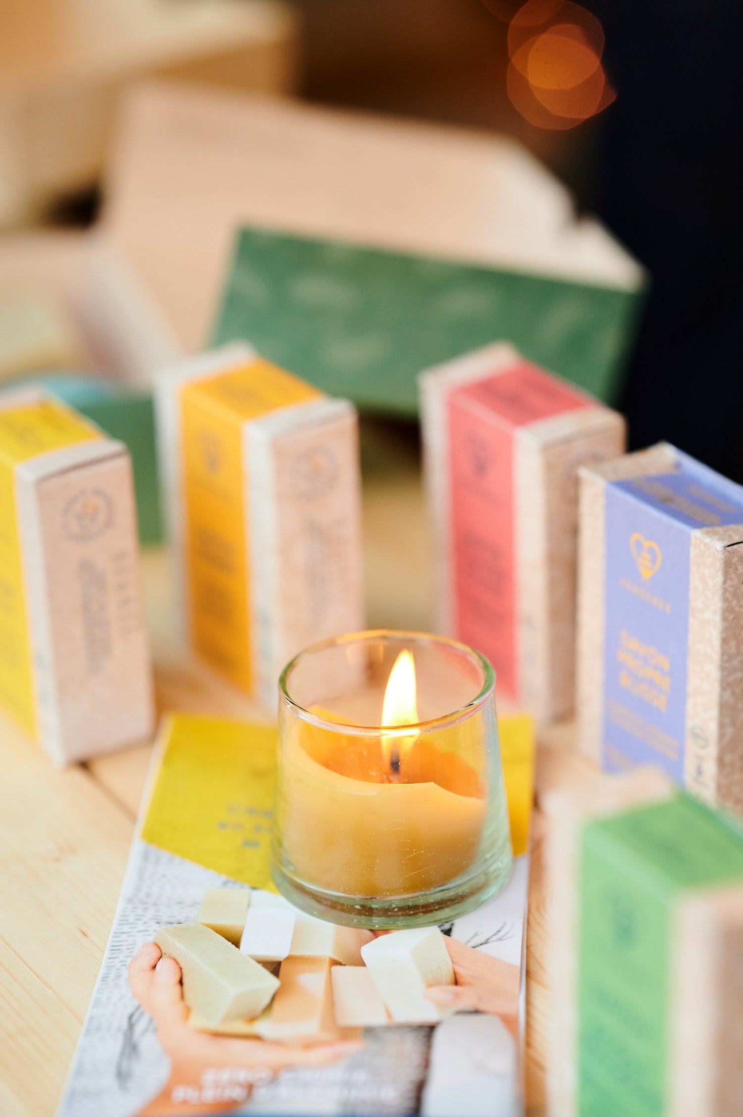 Une bougie en cire d’abeille allumée au centre, entourée de savons Habeebee colorés en boîte et de pépites de savon sur une table en bois.Een brandende bijenwaskaars omringd door kleurrijke Habeebee-zeepdoosjes en zeepblokjes op een houten tafel.