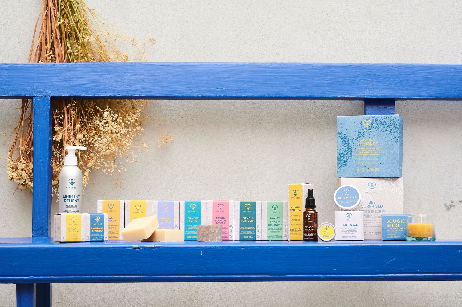 Vue d’ensemble de la gamme de cosmétiques Habeebee, posée sur une étagère bleue : savons solides, huiles, bougies, baumes, shampooings et coffrets cadeaux durables, avec des emballages colorés et naturels.