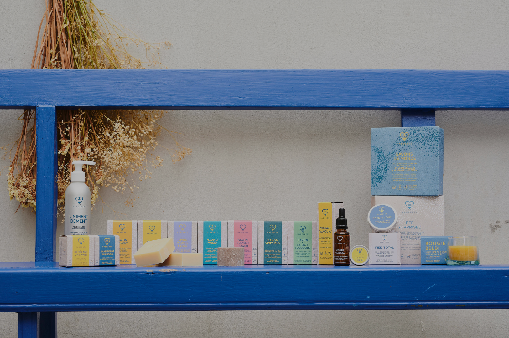 Vue d’ensemble de la gamme de cosmétiques Habeebee, posée sur une étagère bleue : savons solides, huiles, bougies, baumes, shampooings et coffrets cadeaux durables, avec des emballages colorés et naturels.
Overzicht van het volledige assortiment Habeebee-producten op een blauwe plank: vaste zepen, oliën, kaarsen, balsems, shampoos en duurzame geschenkdozen, allemaal met kleurrijke en natuurlijke verpakkingen.
Full view of Habeebee’s product range displayed on a blue shelf: solid soaps, oils, can