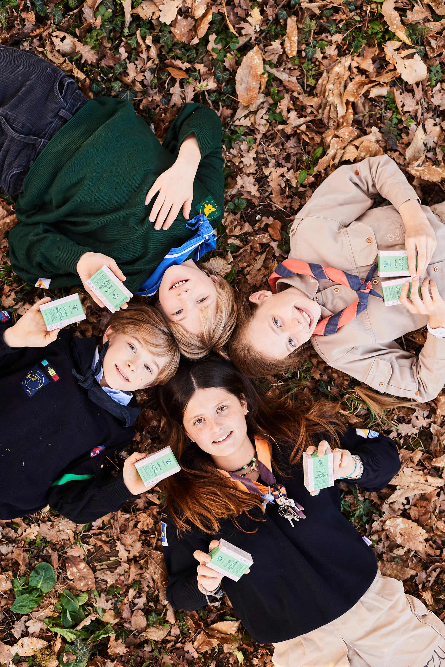 Quatre jeunes scouts allongés dans les feuilles d’automne, souriants et tenant chacun un savon "Scout Toujours" de Habeebee – un savon naturel, artisanal et anti-moustique. Vier jonge scouts liggen in herfstbladeren, lachend met een "Scout Toujours"-zeep van Habeebee in de hand – een natuurlijke, ambachtelijke en muggenwerende zeep. Four young scouts lying in autumn leaves, smiling and holding Habeebee’s "Scout Toujours" soap – a natural, handmade and mosquito-repelling soap.